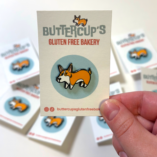 A hand holds a Buttercup's Gluten Free Bakery enamel pin shaped like a corgi on a backing card. The pin features an tan and white corgi inside a light blue circle, set against a blurred background of similar product cards.
