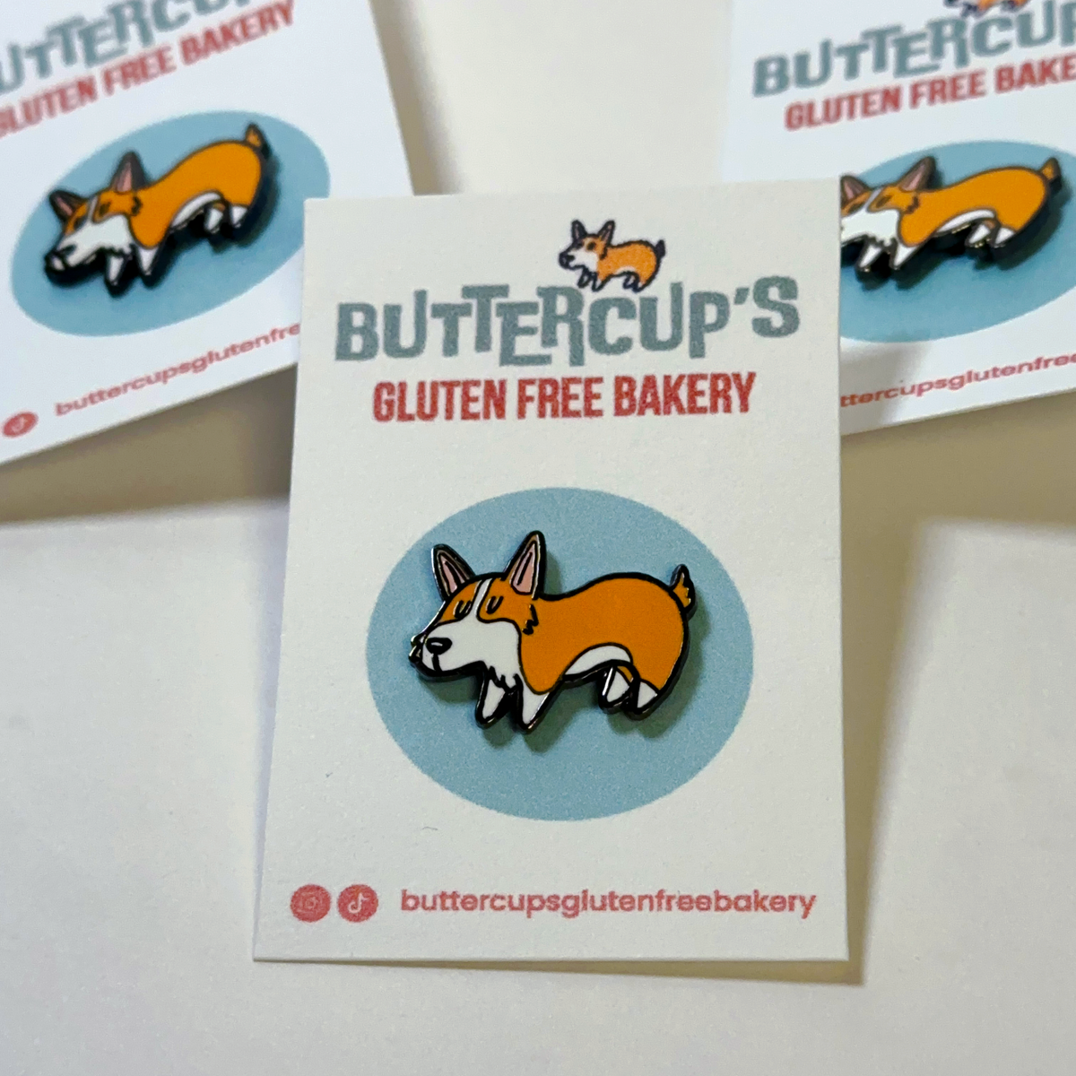 An enamel pin shaped like a cartoon corgi inside a light blue circle, attached to a white backing card that reads 'Buttercup's Gluten Free Bakery.' The card is photographed on a white surface, with two additional copies of the card visible in the background.