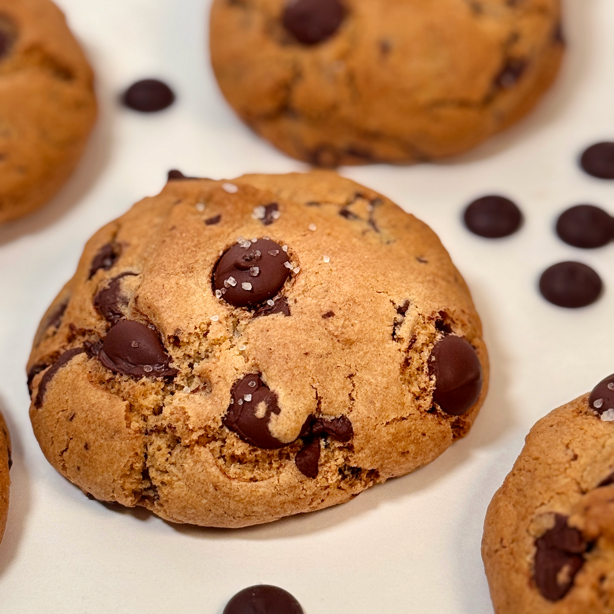 A close-up of a chocolate chip cookie on a white surface, with additional cookies and chocolate chips scattered around it.