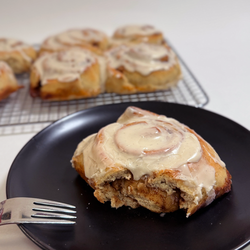 A close-up of a cinnamon roll on a black plate with a fork, with more cinnamon rolls cooling on a wire rack in the background.