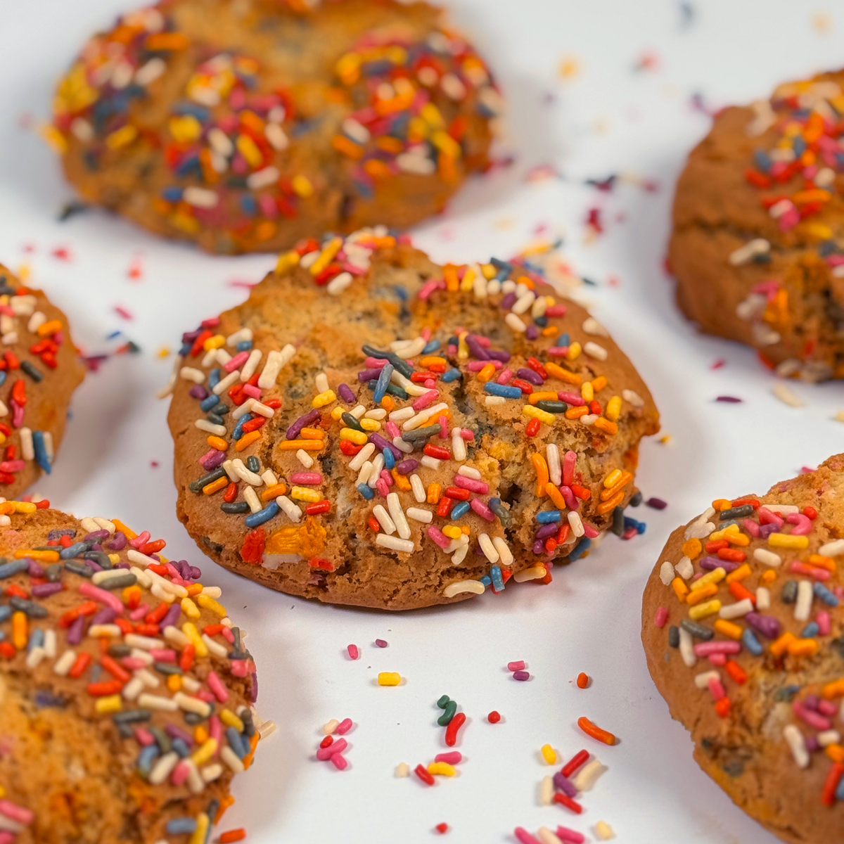 Several golden-brown cookies topped with colorful sprinkles on a white surface, with some sprinkles scattered around.