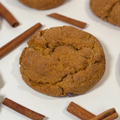 A close-up of a pumpkin snickerdoodle 
cookie with sugar dusting, surrounded by scattered cinnamon sticks on a white surface.
