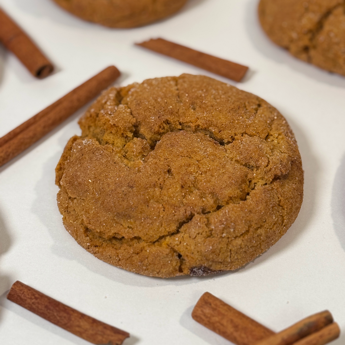 A close-up of a pumpkin snickerdoodle 
cookie with sugar dusting, surrounded by scattered cinnamon sticks on a white surface.