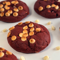 A close-up of a round red velvet cookie topped with small white chocolate chips, with additional cookies and scattered white chocolate chips on a white surface.