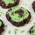 Chocolate cookies topped with mint white chocolate and crushed dark cookie crumbs, arranged on a white surface with a few scattered mint chocolate chips.